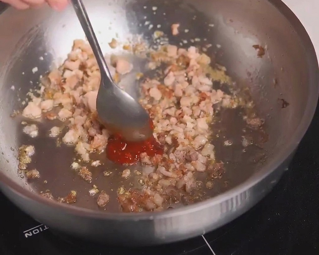 Une cuillère en métal ajoutant de la pâte de haricots rouge pimentée à la viande hachée dorée dans la poêle.