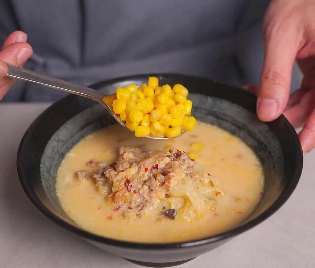 Une cuillère déposant une portion de maïs jaune croquant sur le dessus dun bol de ramen fumant.