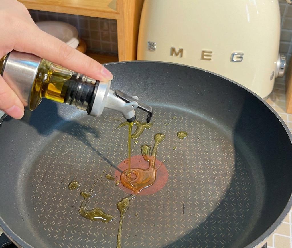 De lhuile de cuisson versée depuis un flacon en verre dans une poêle antiadhésive noire sur un plan de travail.