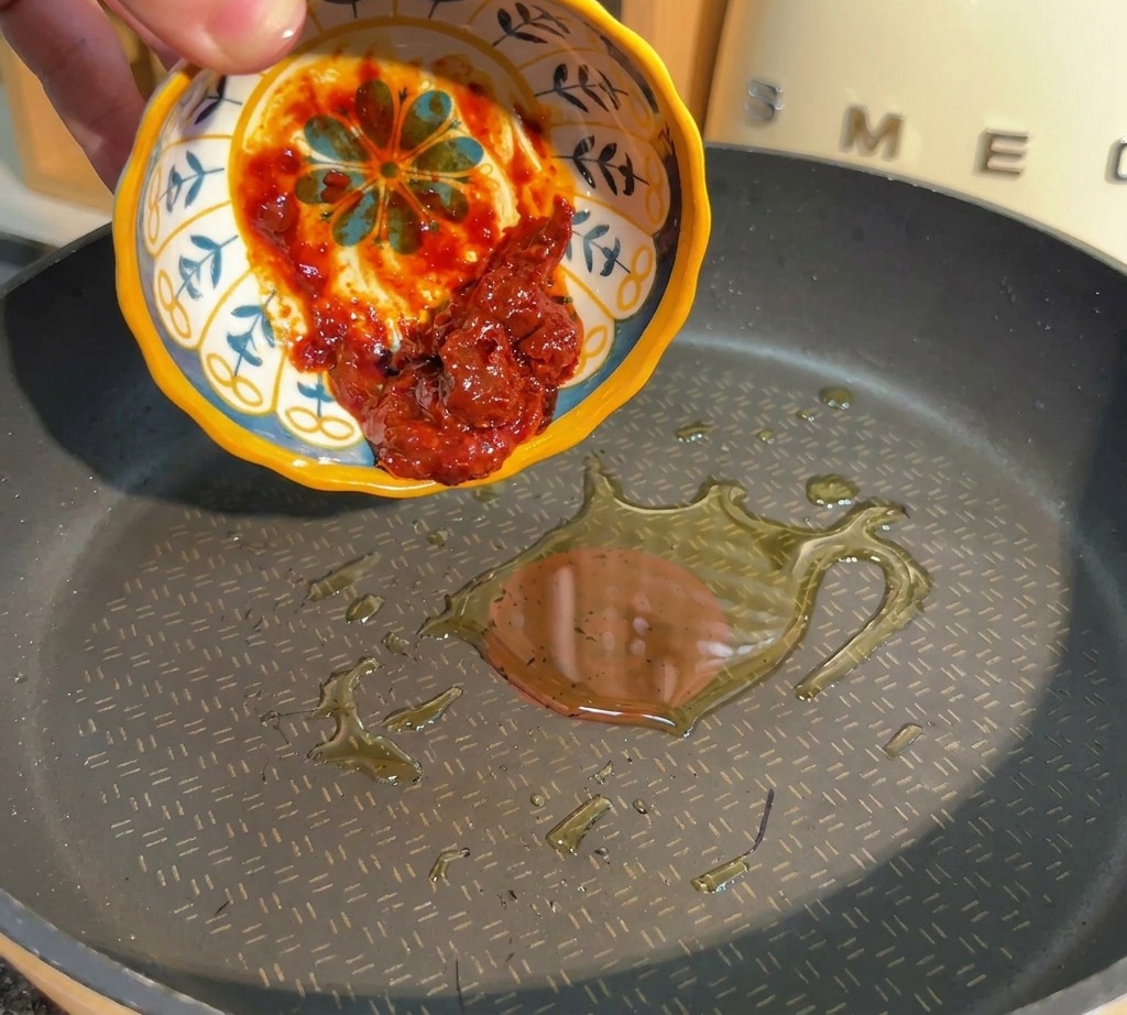 Une main versant de la pâte de piment rouge depuis un petit bol vers une poêle contenant de lhuile chaude.