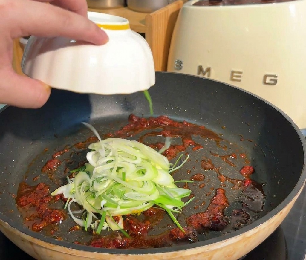 Des oignons verts émincés ajoutés depuis un bol blanc dans une poêle contenant la base de sauce rouge épicée.
