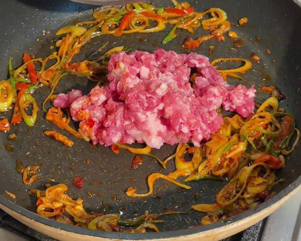 Du porc haché cru déposé dans une poêle contenant déjà les oignons verts sautés et la pâte de piment rouge.