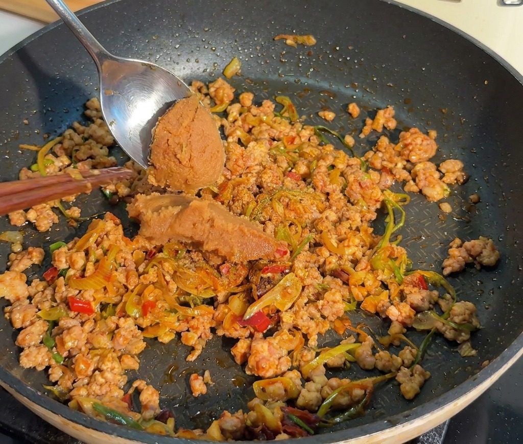 Une cuillère en métal ajoutant une portion de pâte de miso brune épaisse dans une poêle de viande hachée cuite.