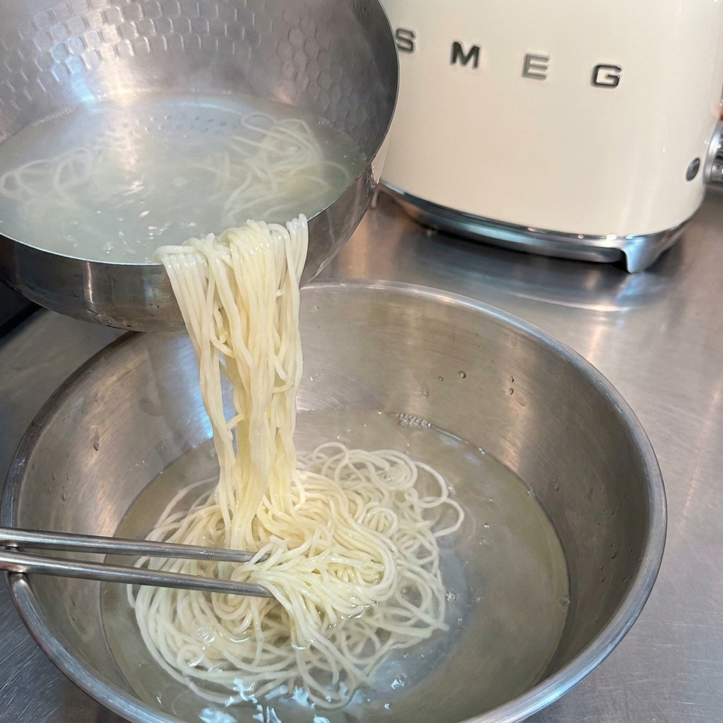 Nouilles ramen cuites rincées et remuées avec des baguettes en métal dans un bol en inox rempli deau froide.