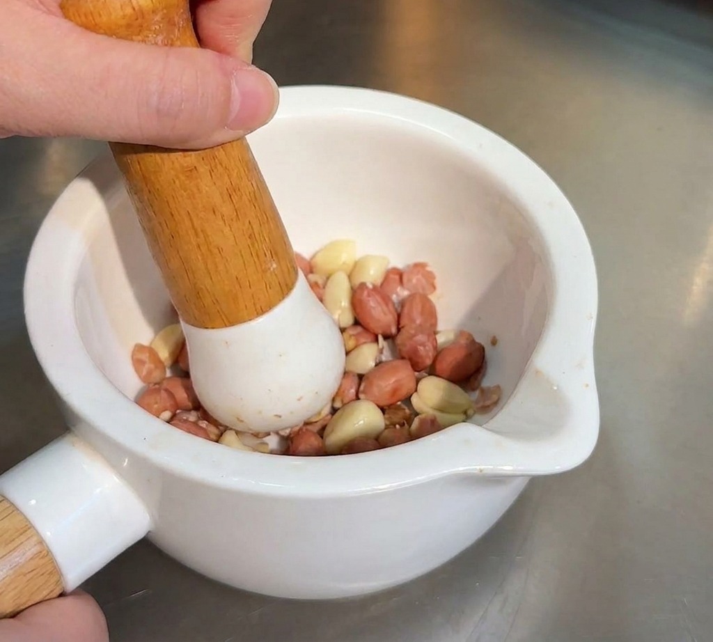 Une personne utilisant un pilon en bois pour écraser des cacahuètes grillées et des graines de sésame dans un mortier en céramique blanc.