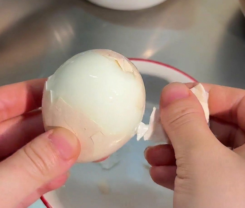Des mains écalant soigneusement la coquille dun œuf mollet blanc et lisse au-dessus dun petit bol.