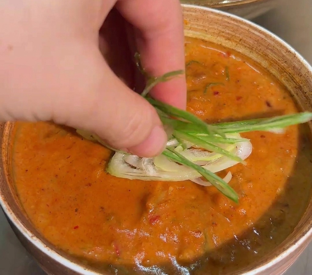 Une main déposant un nid de fines lamelles doignons verts sur la surface dune sauce épaisse orange dans un bol en céramique.