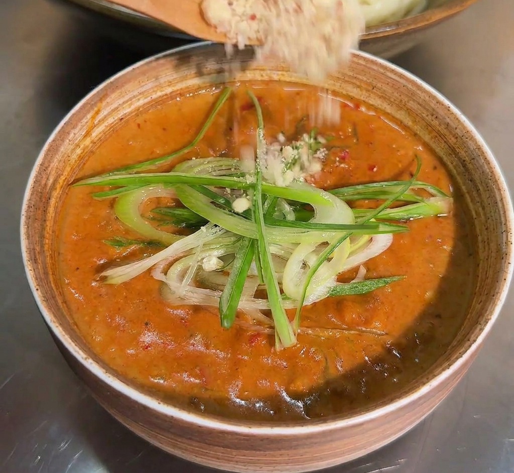 Une cuillère en bois saupoudrant un mélange de noix et graines broyées sur un bol de sauce déjà garni doignons verts.