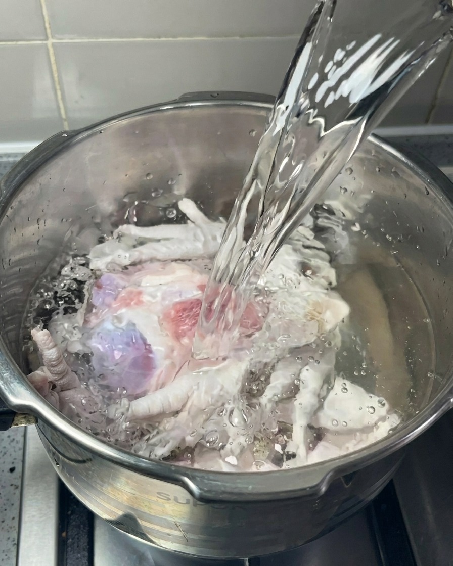 Un filet deau claire versé dans une marmite contenant des os de porc et des pattes de poulet.