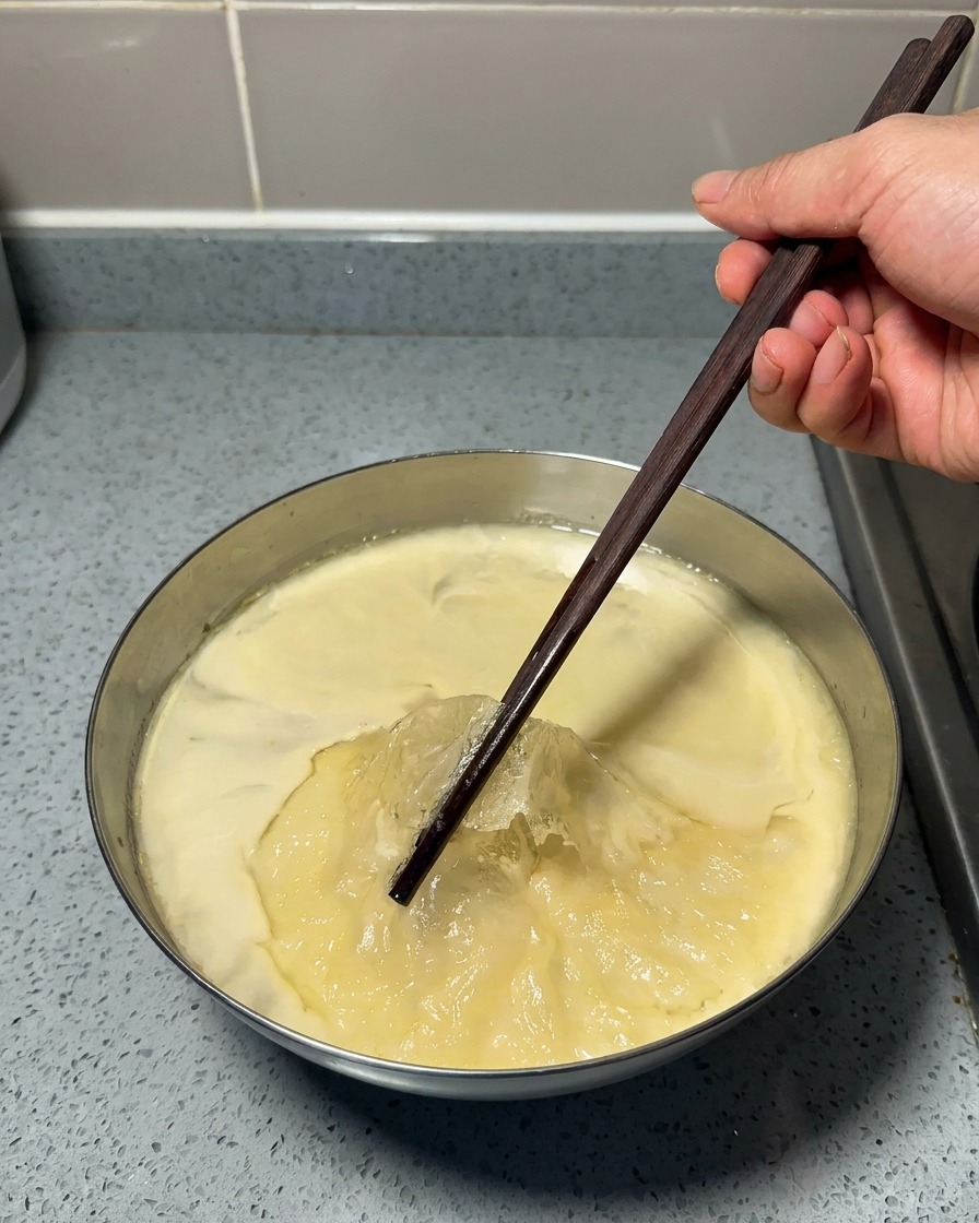 Une main utilisant des baguettes en bois pour retirer une fine couche de graisse jaune sur un bouillon gélatineux refroidi.