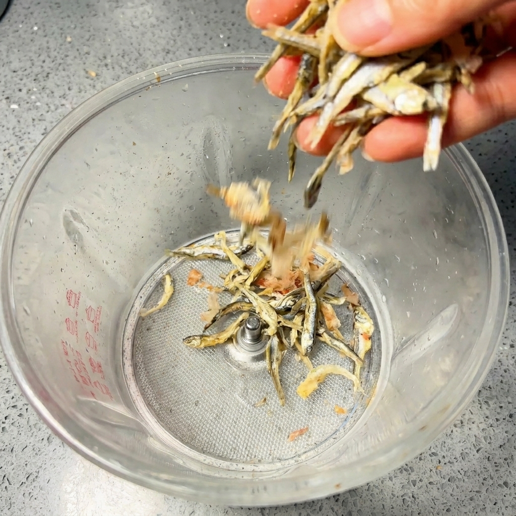 Une main versant des anchois séchés et des flocons de bonite dans le bol dun mixeur transparent.