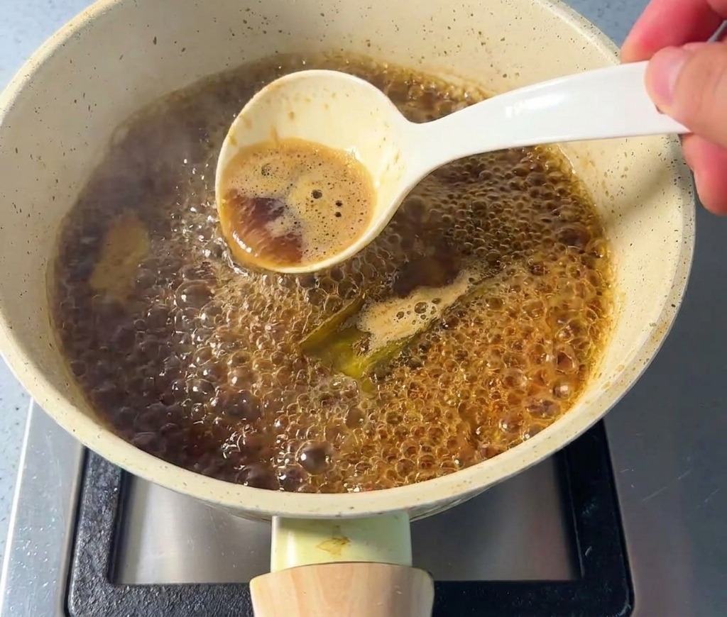 Une main tenant une cuillère blanche pour retirer lécume à la surface dune sauce brune bouillonnante.
