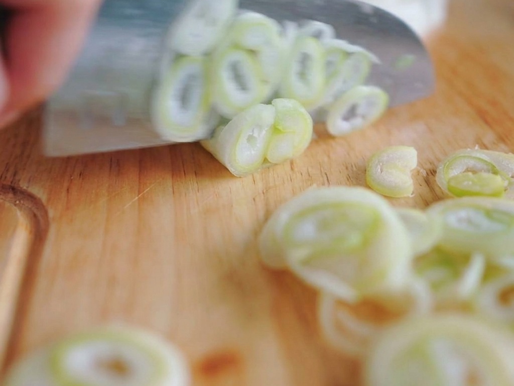 Couteau de chef découpant des tiges doignons verts en fines rondelles sur une planche à découper.