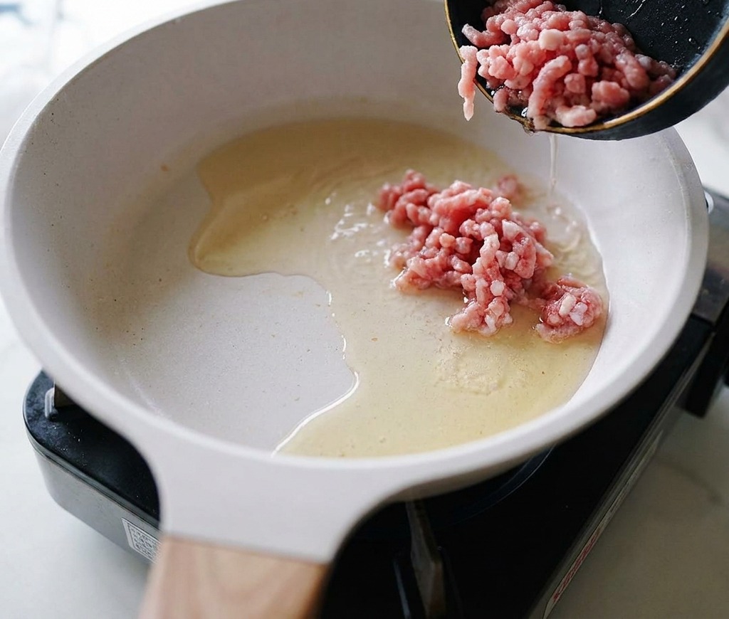 Porc haché cru versé dune poêle noire dans une poêle en céramique blanche avec de lhuile chaude.