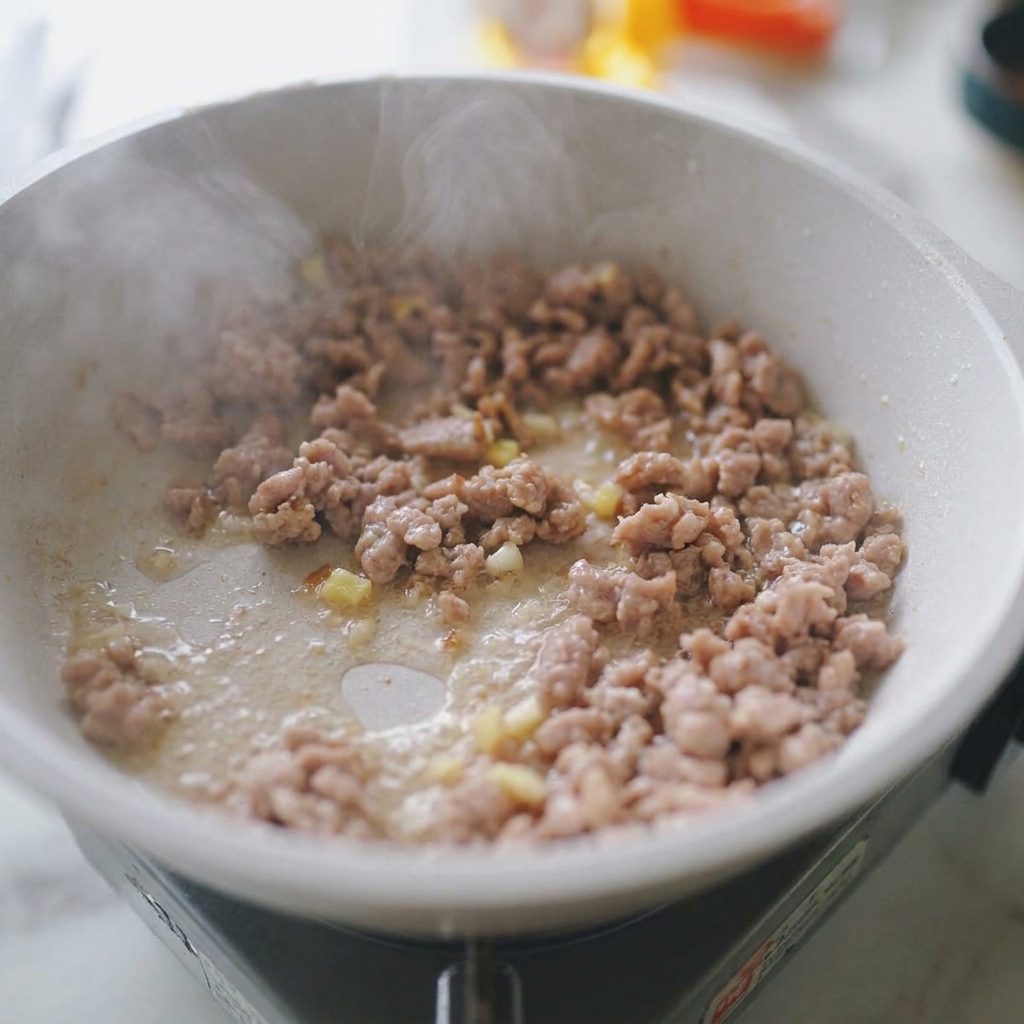 Porc haché sautant dans une poêle blanche avec de lail et du gingembre hachés visibles.