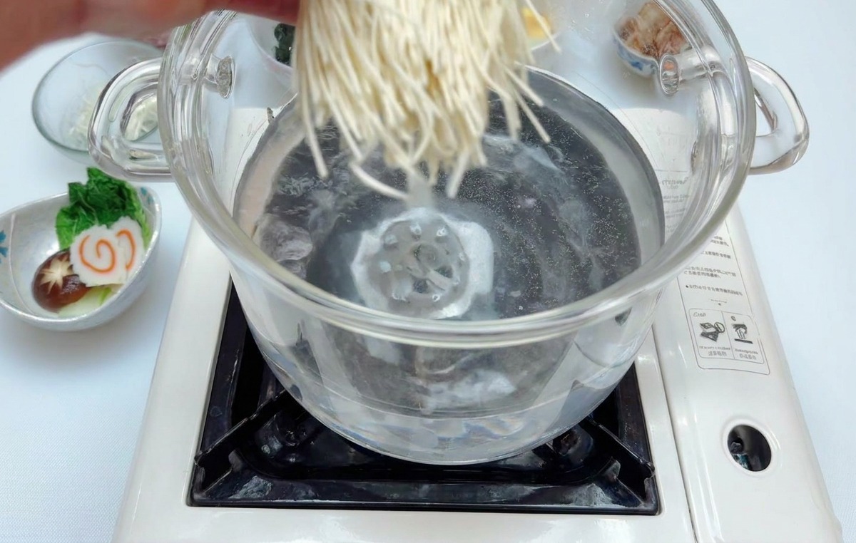 Une main déposant des nouilles ramen blanches dans une casserole deau bouillante.