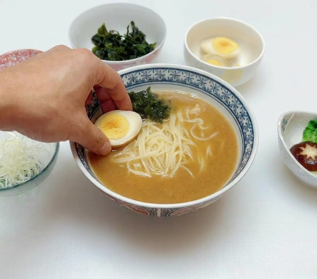 Une main plaçant un demi-œuf mollet sur un bol de ramen contenant déjà le bouillon et le wakame.