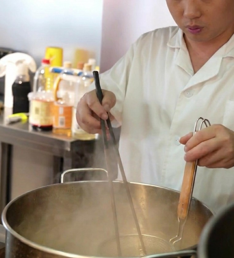 Un chef plongeant une passoire à ramen remplie de nouilles fraîches dans une grande marmite deau bouillante.