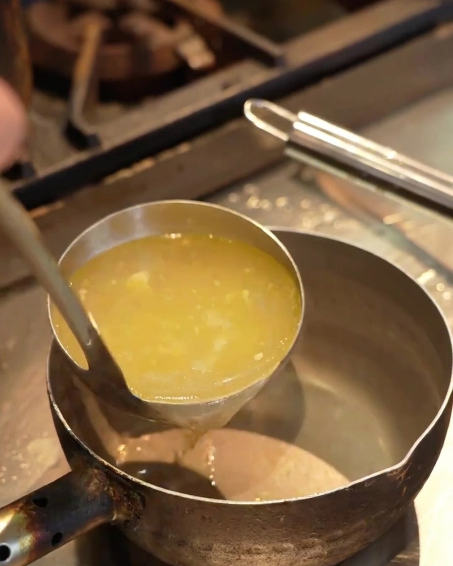 Bouillon jaune clair versé dune louche dans une petite casserole de service sur un plan de travail.