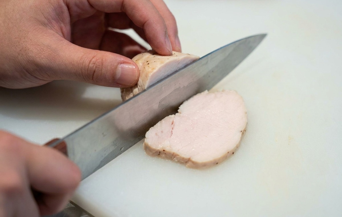 Mains tranchant un morceau de blanc de poulet tendre en rondelles fines sur une planche à découper blanche.