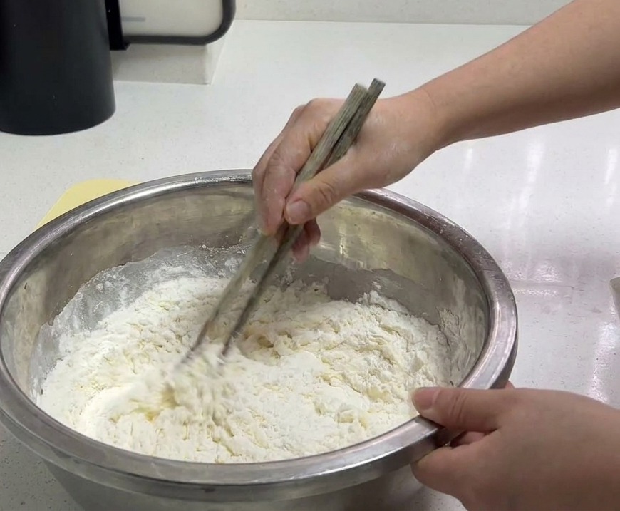 Une personne utilisant des baguettes en bois pour mélanger la farine et leau dans un bol en inox afin de former une pâte grumeleuse.