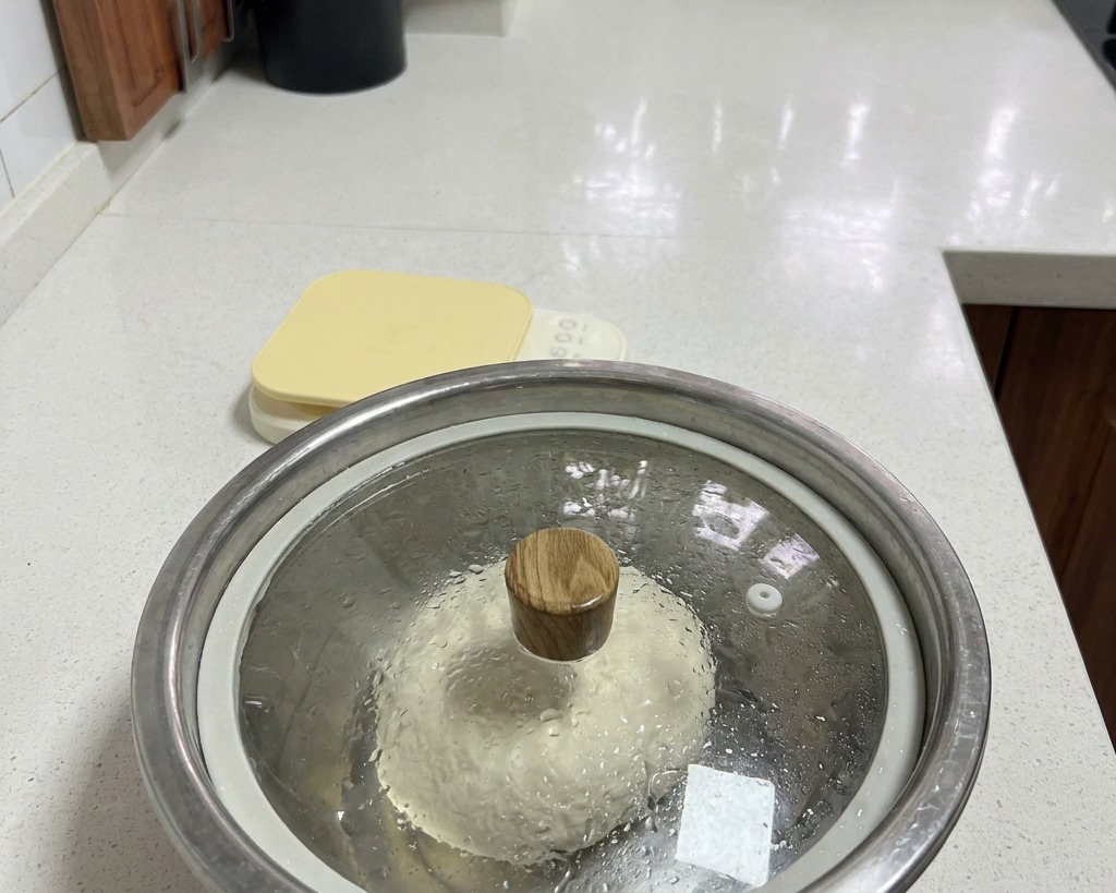 Un bol en inox contenant la pâte, recouvert dun couvercle en verre avec de la condensation visible à lintérieur.
