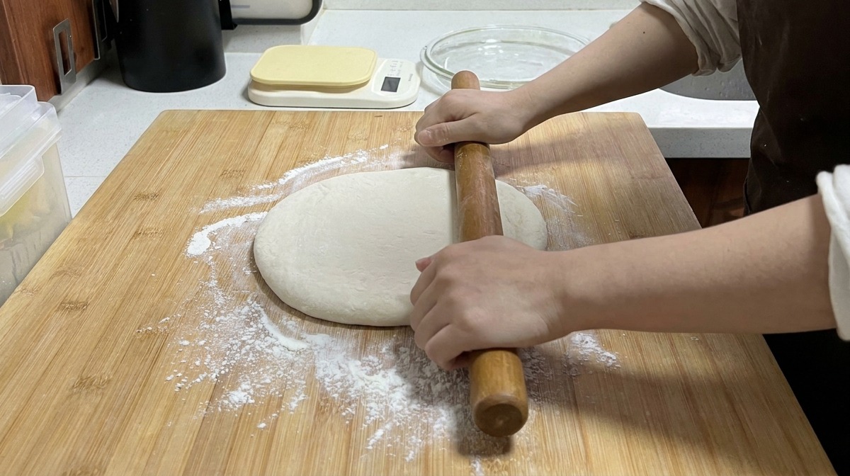 Mains utilisant un long rouleau à pâtisserie en bois pour aplatir une boule de pâte sur une planche saupoudrée de farine.