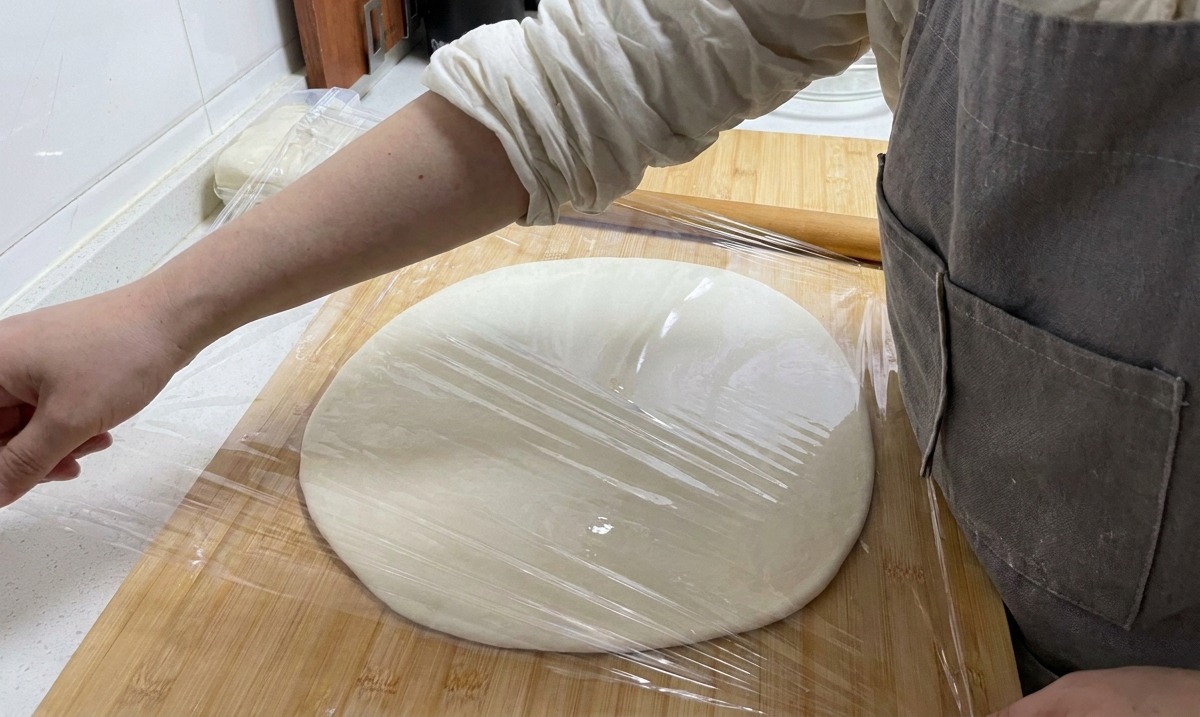 Une main recouvrant la feuille de pâte étalée avec du film plastique transparent sur une planche en bois.