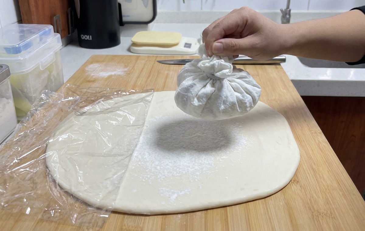 Une main secouant un petit sachet en tissu pour libérer un voile de farine blanche sur la pâte étalée.