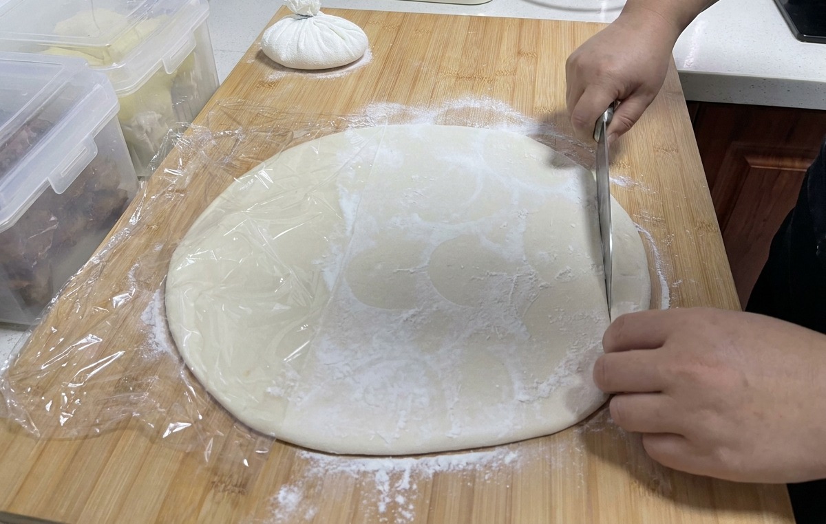 Un grand couteau en inox découpant une feuille de pâte blanche sur une planche en bois pour créer de longues bandes.