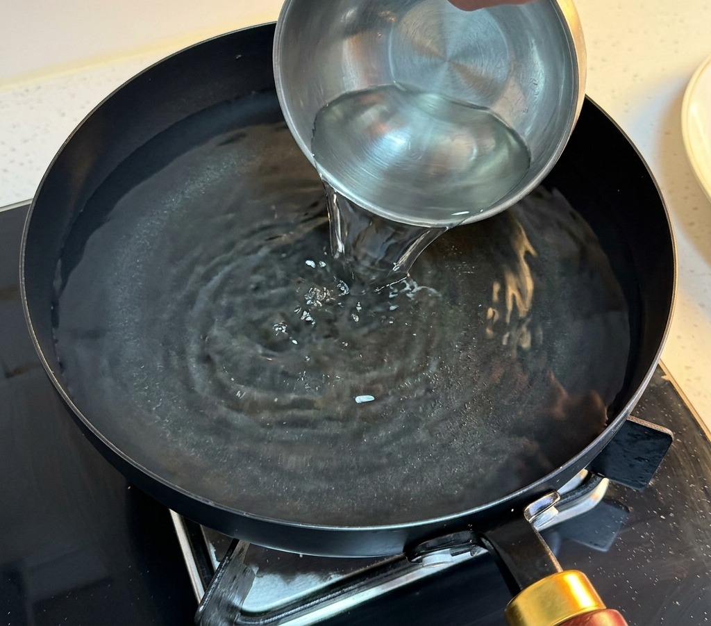 Une main versant de leau claire dun petit bol en métal dans une poêle remplie deau chaude.