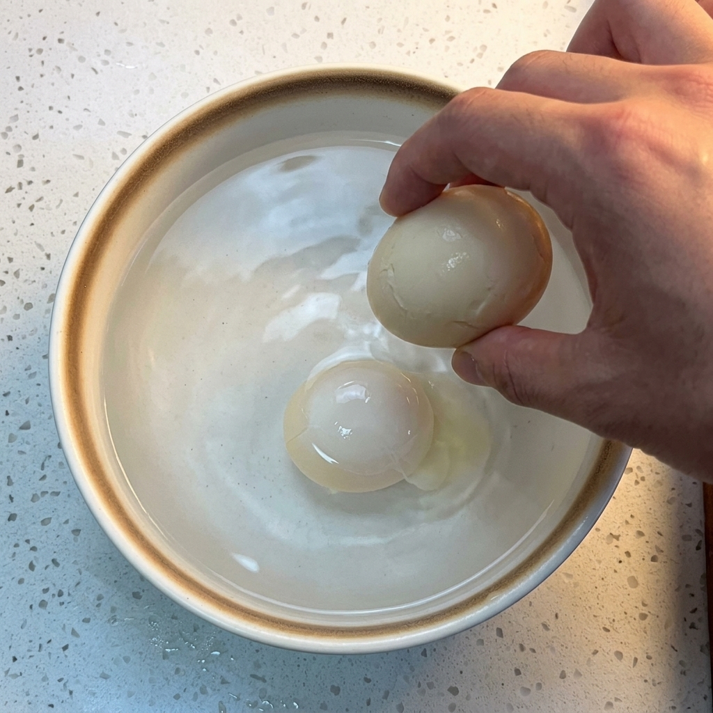 Un œuf cuit plongé dans un bol en céramique blanche rempli deau froide et de glaçons.