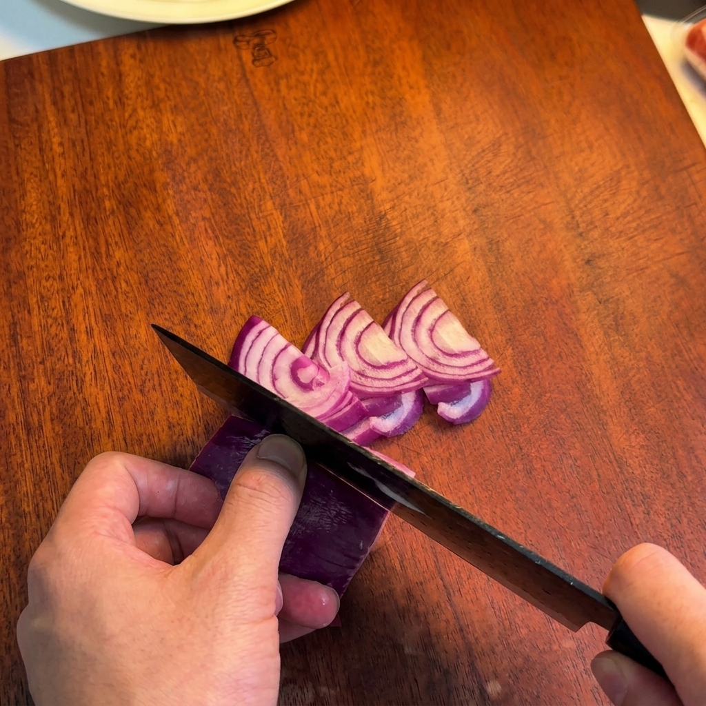 Une personne utilisant un couteau de chef pour couper un oignon rouge en fines lamelles sur une planche en bois.