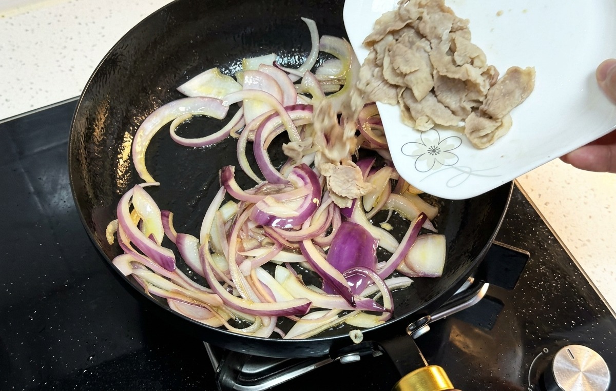 Tranches de bœuf cuites versées dune assiette blanche dans une poêle contenant des oignons rouges sautés.
