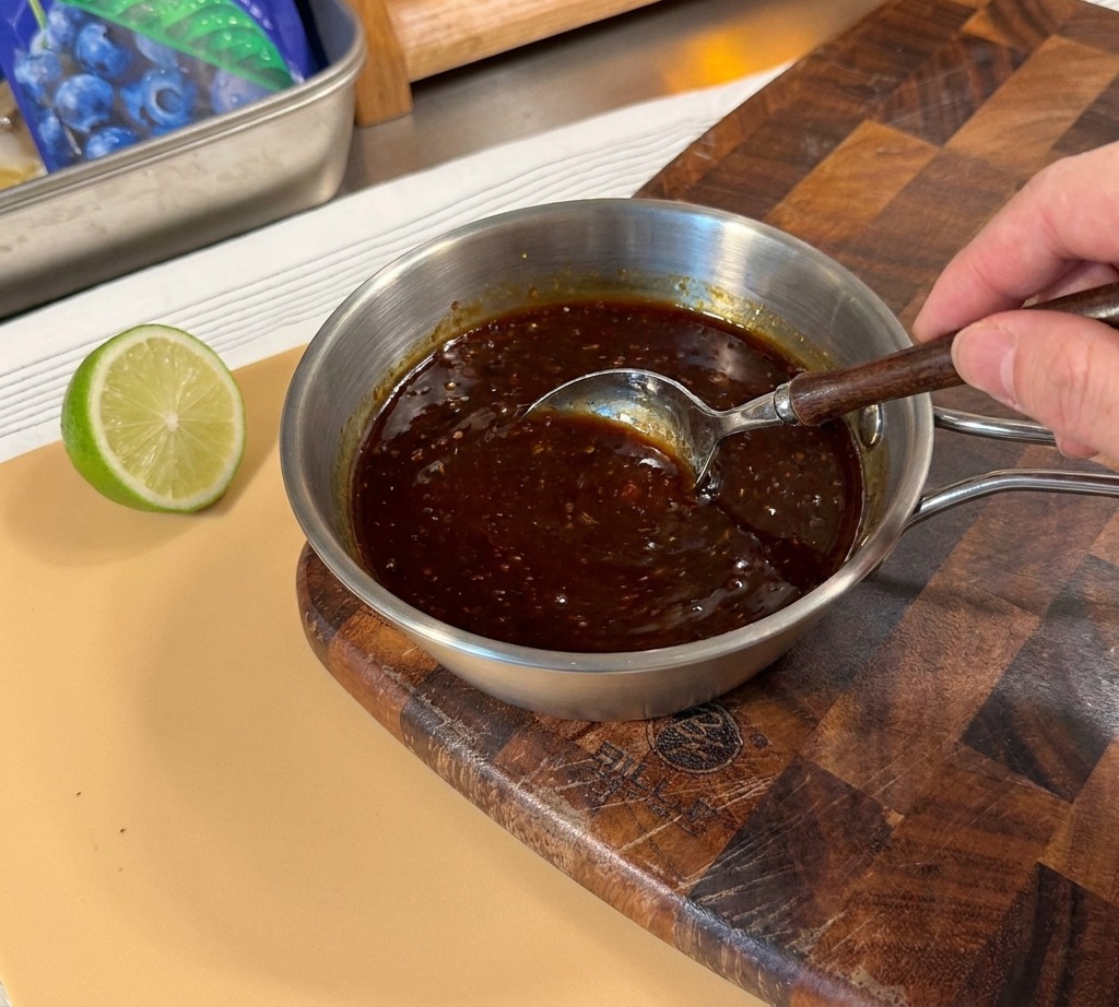Une main utilisant une cuillère pour mélanger une sauce teriyaki épaisse et brune dans un petit pot en métal.