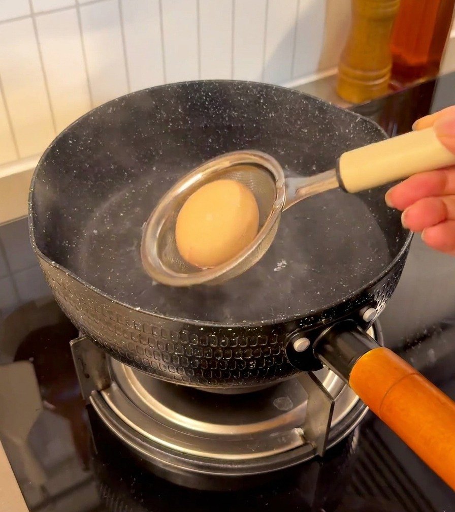 Un œuf dans une petite passoire en filet plongé dans une casserole deau fumante sur une cuisinière.