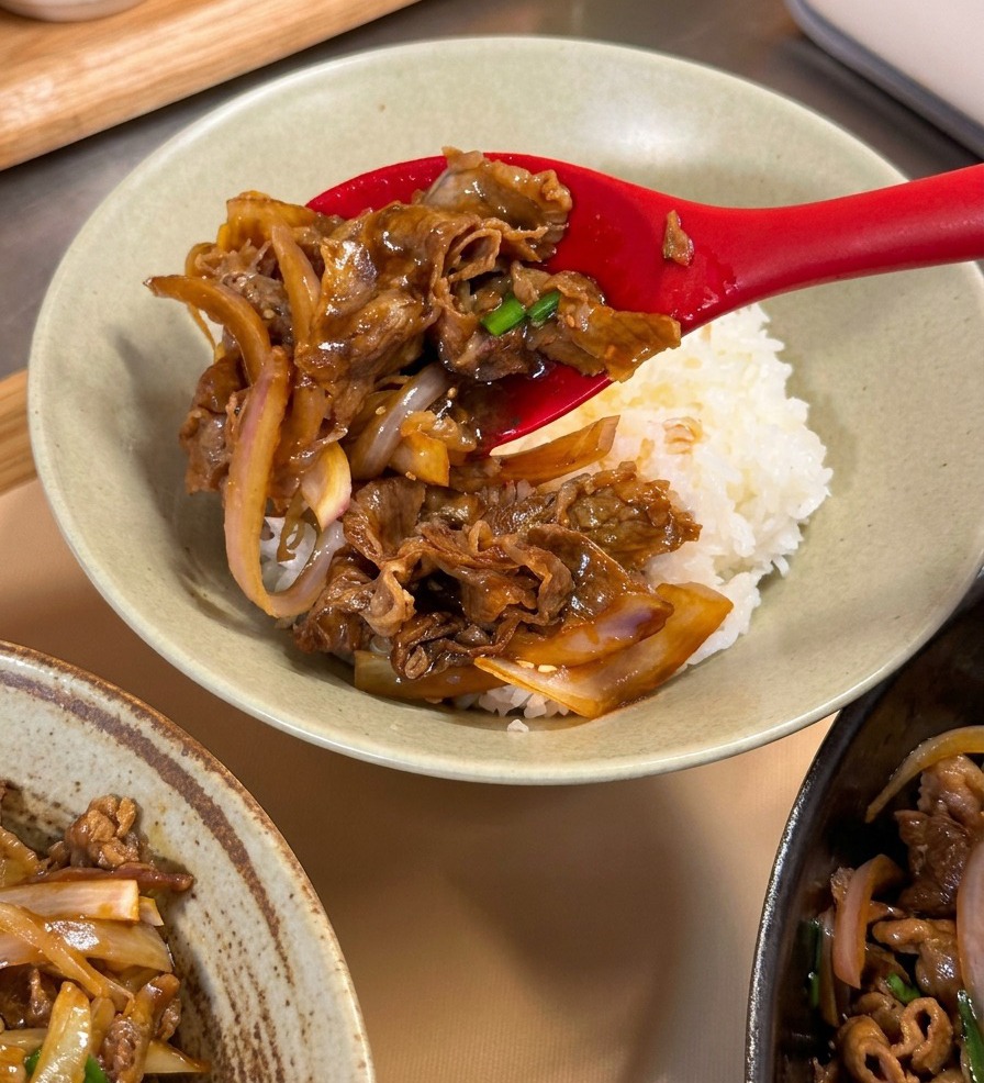Une cuillère rouge déposant une portion de bœuf teriyaki brillant sur un lit de riz blanc dans un bol en céramique.