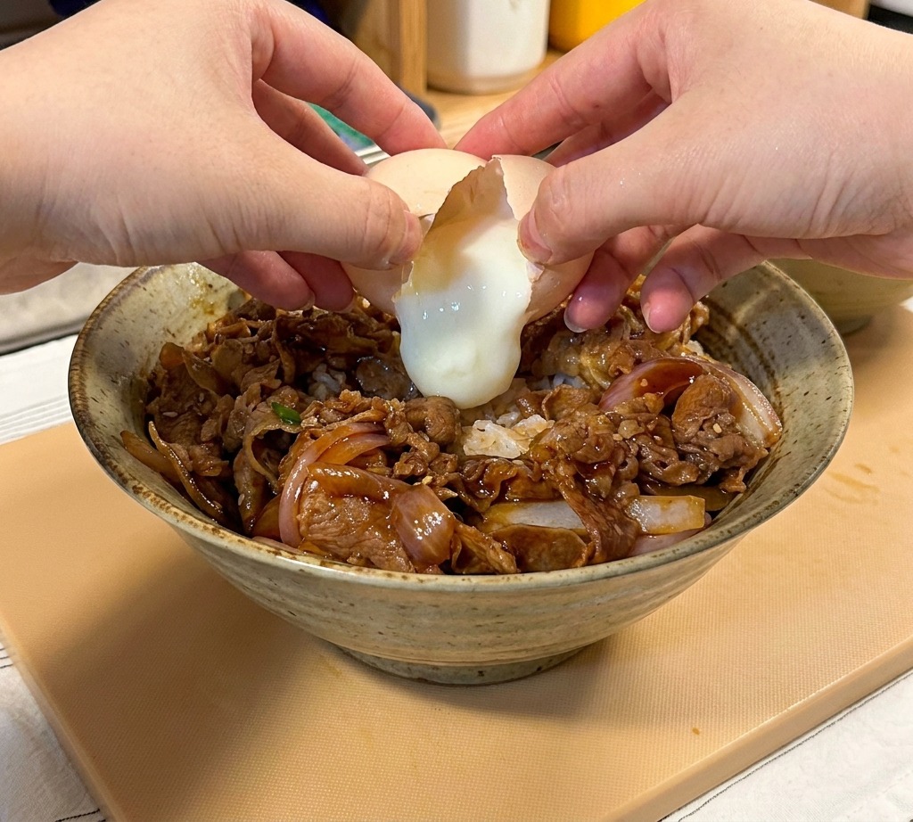 Gros plan de deux mains cassant un œuf onsen mollet sur un bol de bœuf teriyaki servi sur du riz blanc.
