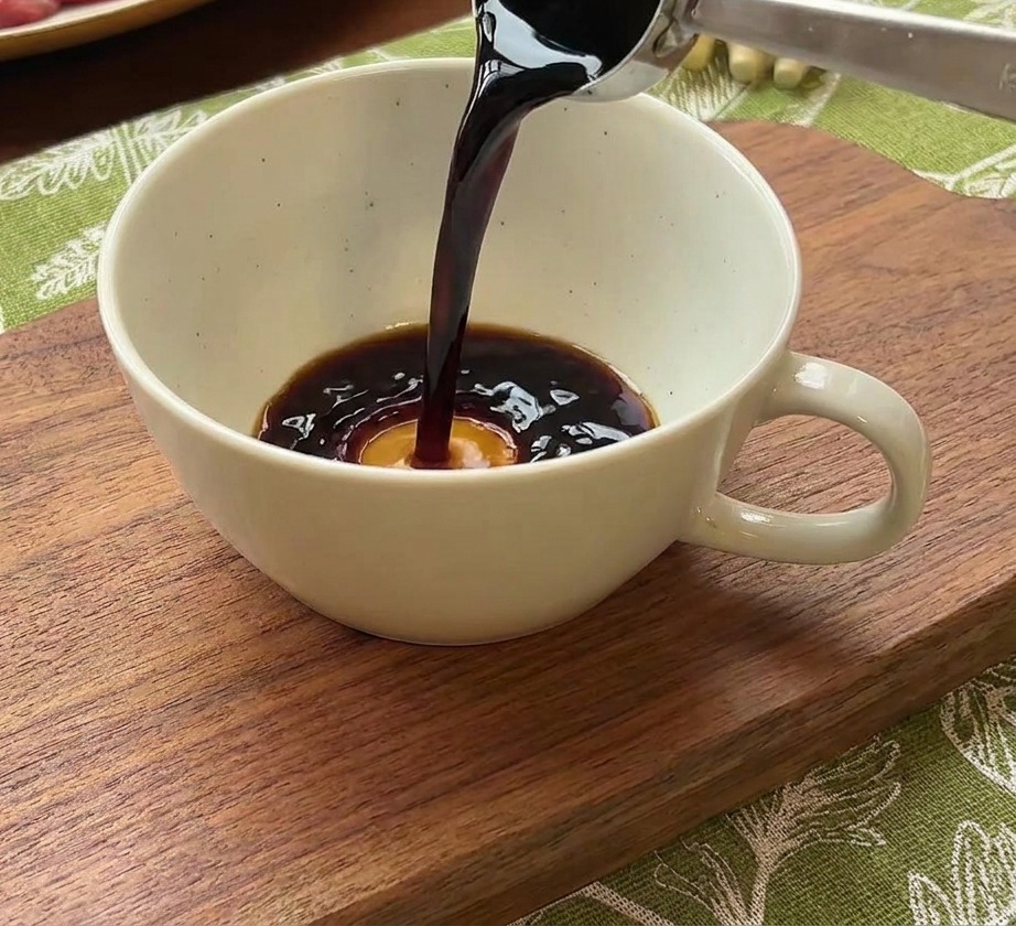 Une cuillère en métal versant de la sauce soja dans un bol contenant de leau et une dose de pâte de piment rouge.