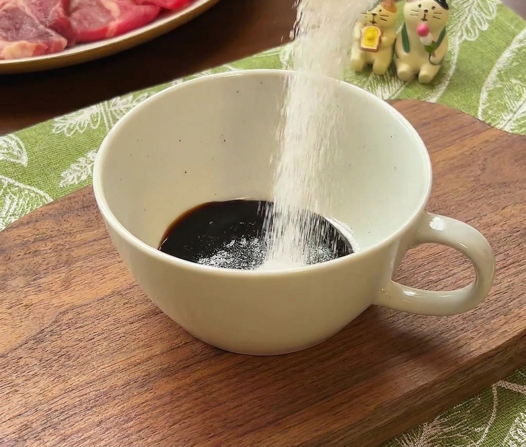 Du sucre blanc versé dune cuillère de mesure dans un bol contenant une sauce sombre.