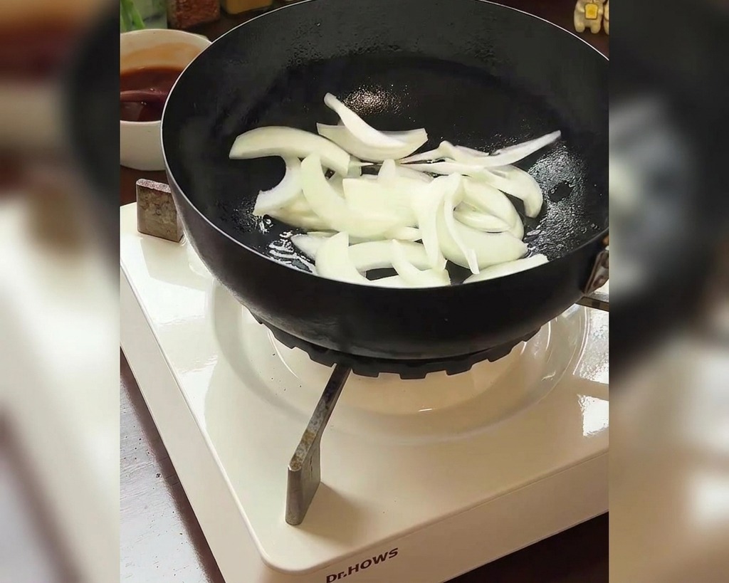 Lanières doignons blancs ajoutées dans une poêle avec un bol de kimchi rouge visible en arrière-plan.