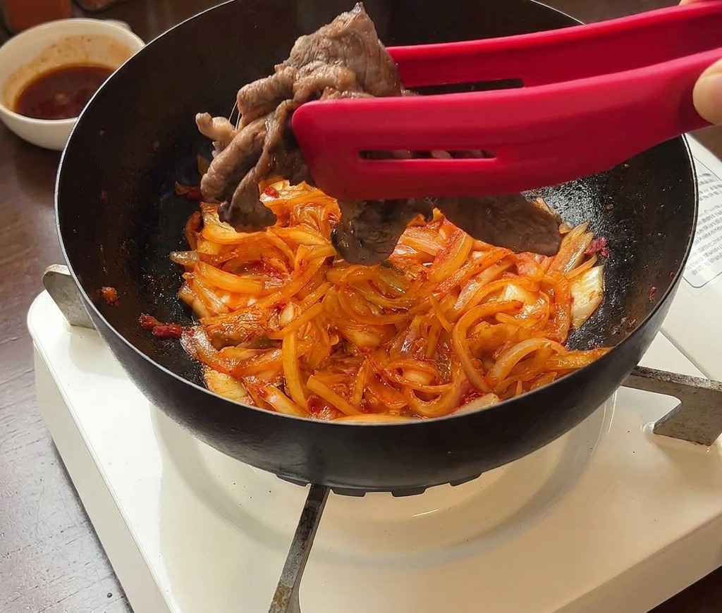 Tranches de bœuf saisies ajoutées dans une poêle contenant du kimchi et des oignons à laide de pinces rouges.