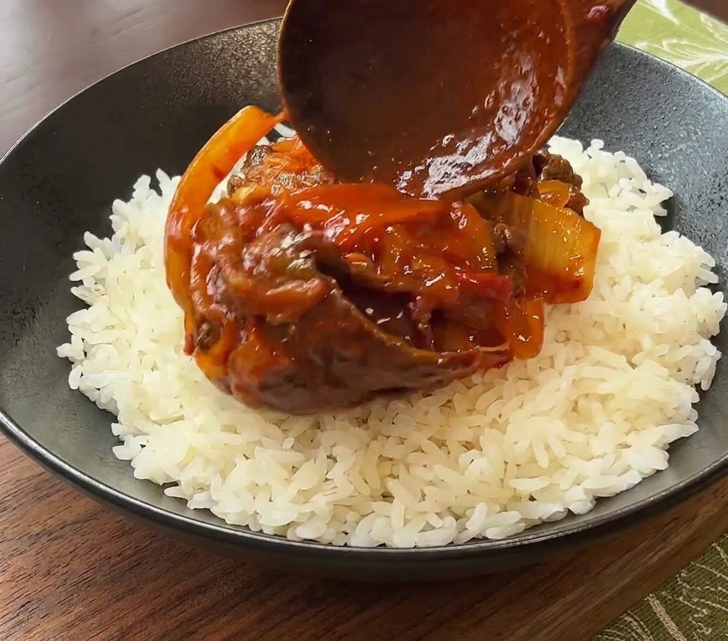 Une cuillère en bois déposant une portion de bœuf et kimchi en sauce sur un lit de riz blanc dans un bol noir.