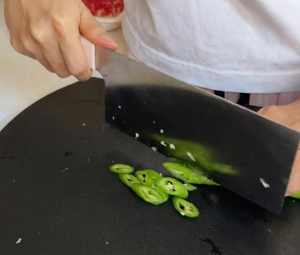 Un couperet de cuisine tranchant des piments verts frais sur une planche à découper noire.
