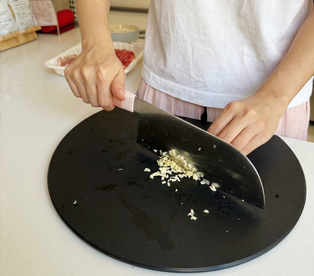 Gros plan de mains hachant finement des gousses dail sur une planche à découper noire.