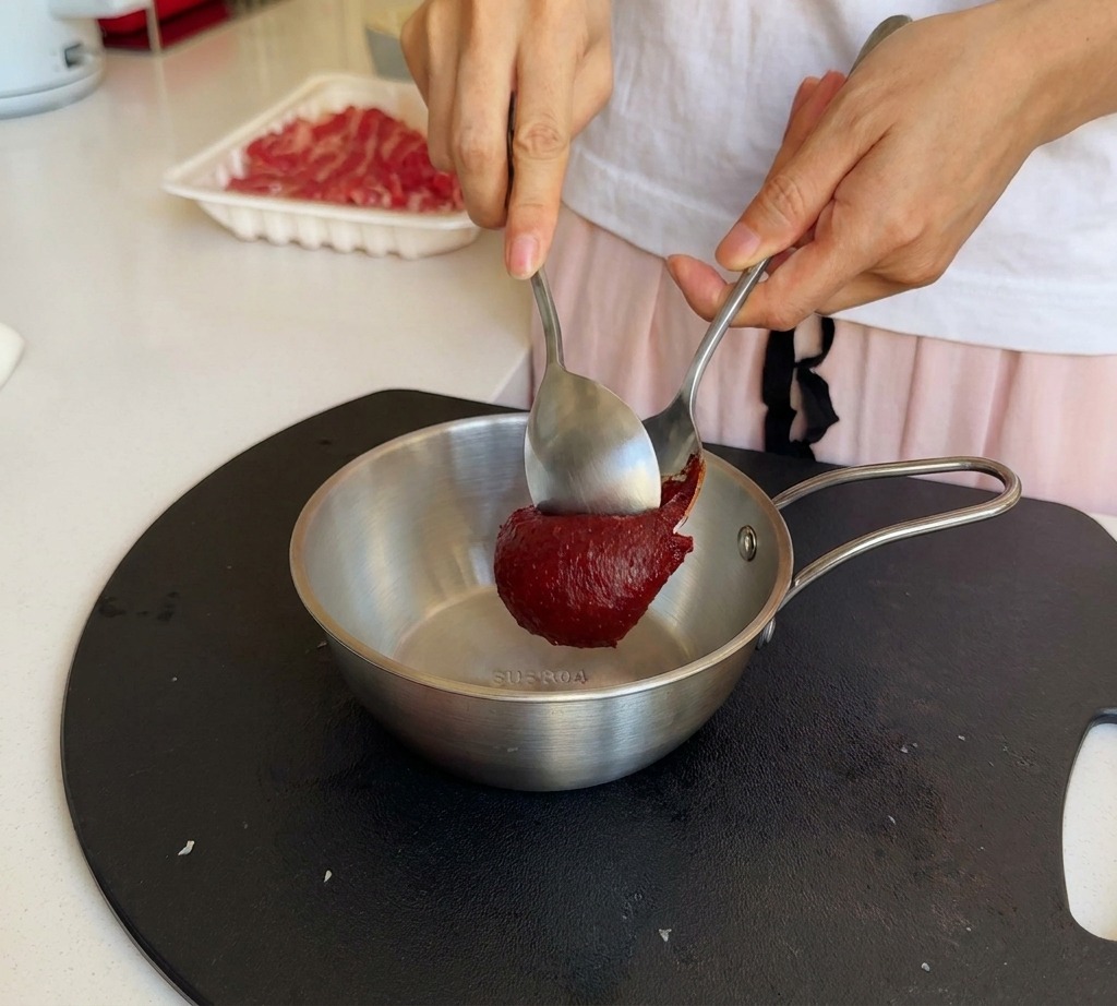 Utilisation de deux cuillères en métal pour ajouter de la pâte de piment coréenne rouge dans un bol en inox.