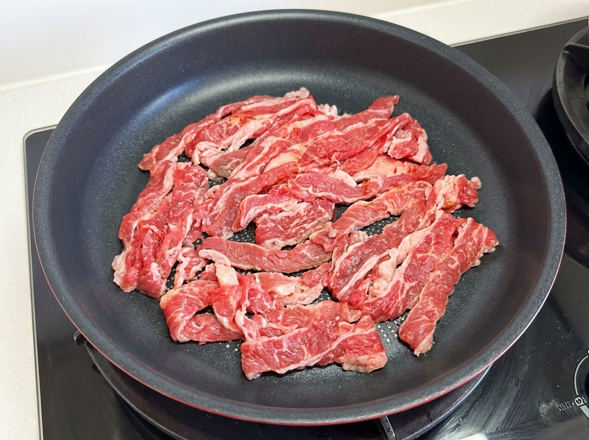 Fines tranches de bœuf gras cru étalées dans une poêle antiadhésive noire sur le feu.
