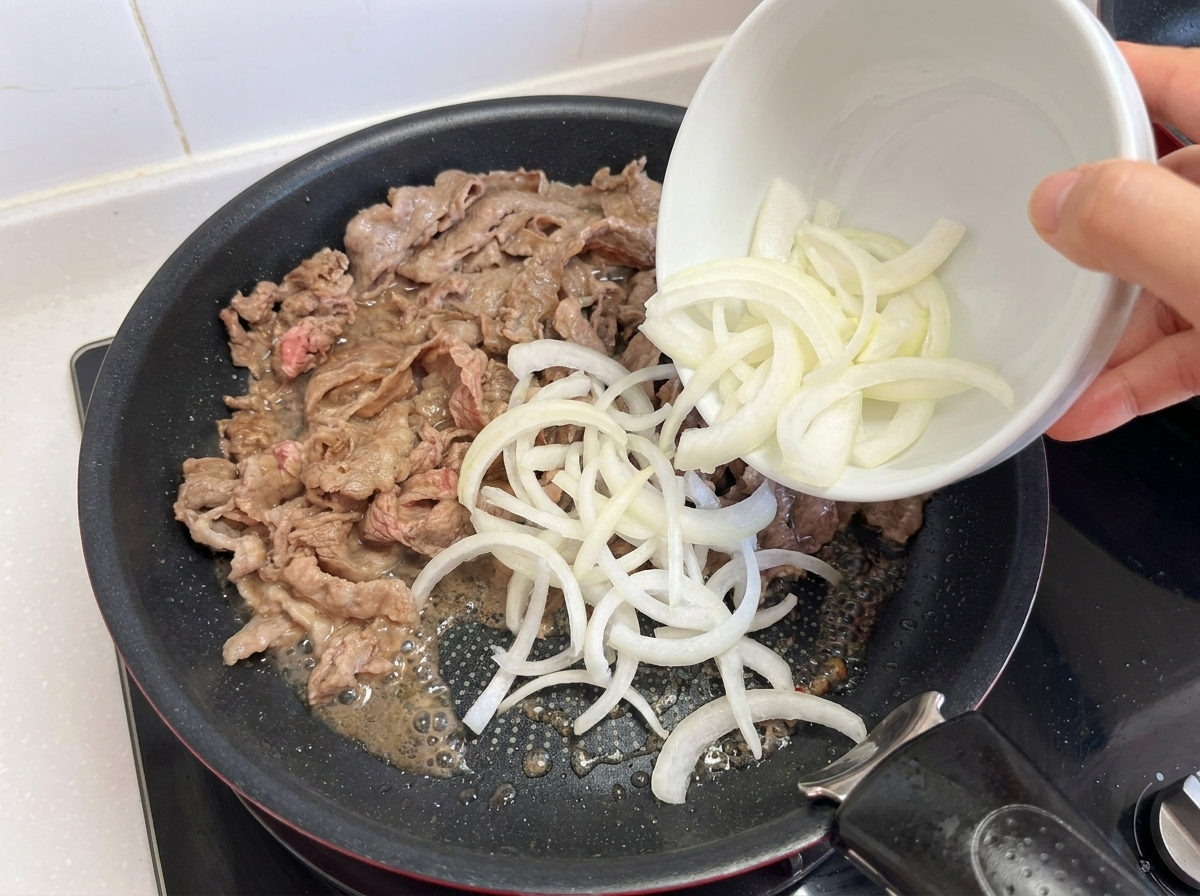 Une main versant un bol doignons émincés dans une poêle contenant du bœuf partiellement cuit.