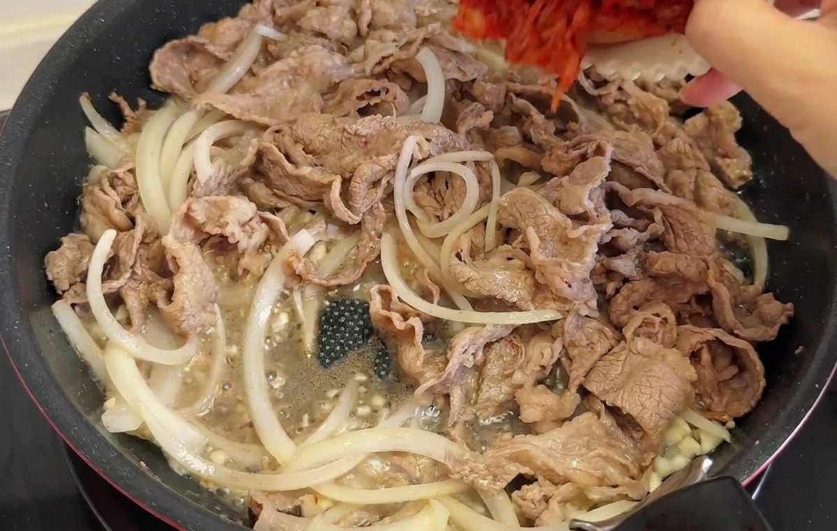 Une main versant du kimchi rouge haché et des piments verts dans une poêle avec le bœuf et les oignons.