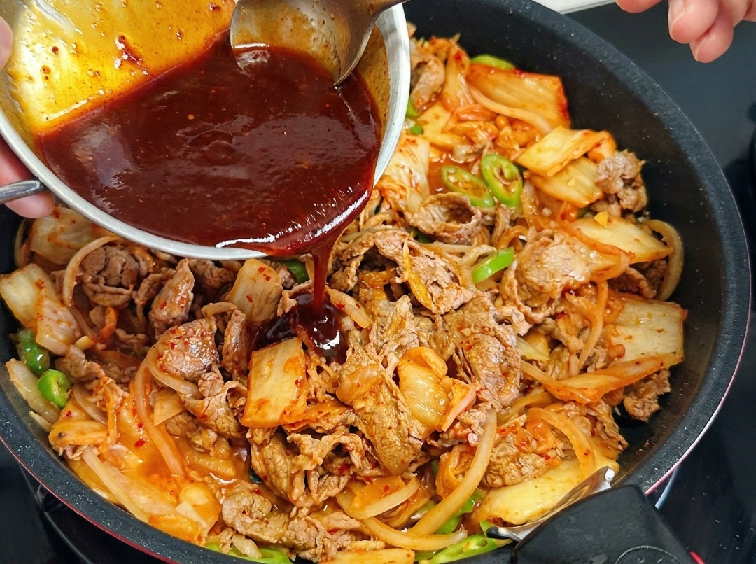 Personne versant une sauce rouge foncé depuis un bol en métal dans une poêle garnie de bœuf, kimchi et piments.